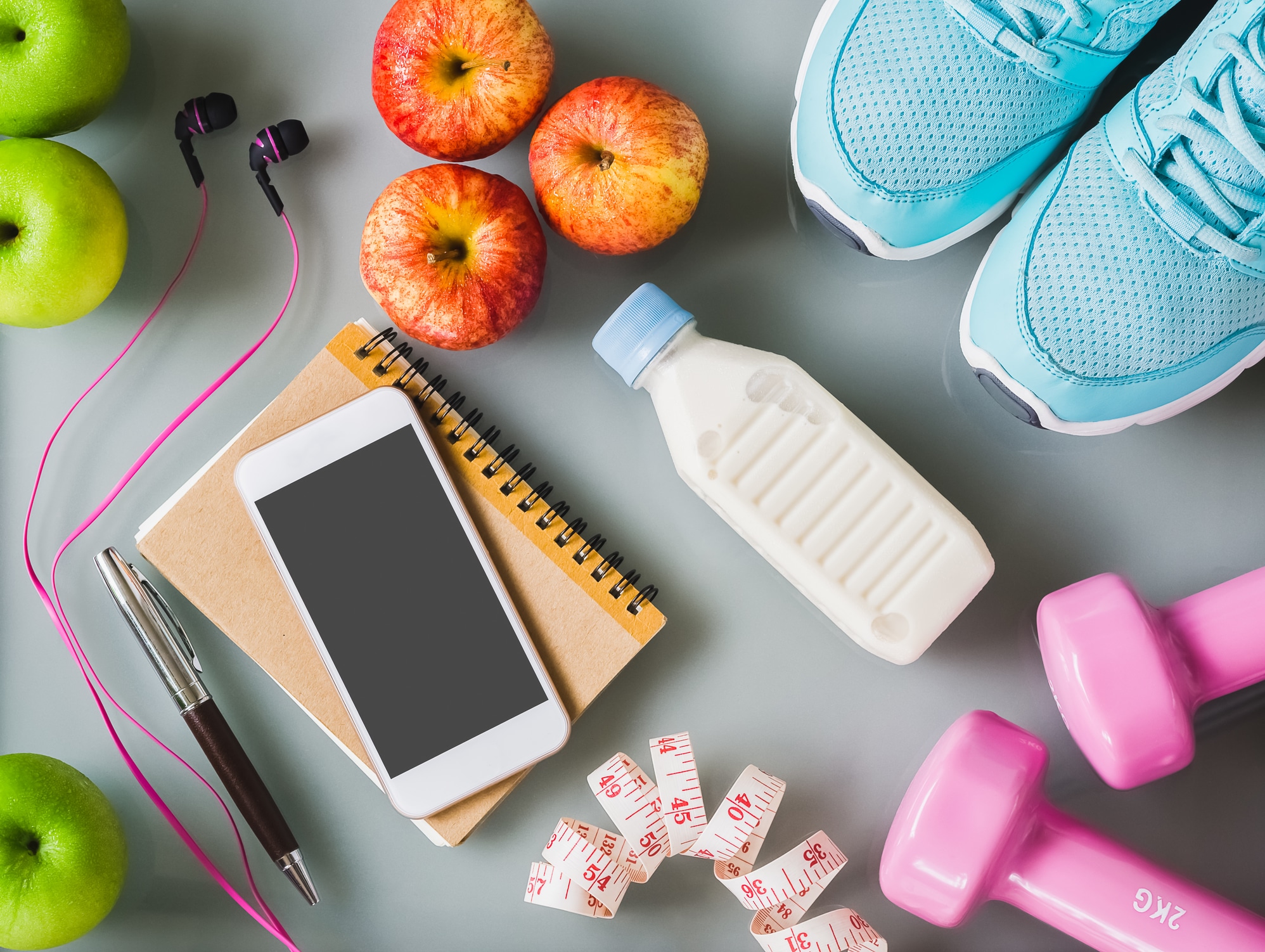 Red and green apples, accompanied by a notebook, a cellphone, headphones, a milk bottle, dumbbells, and exercise tennis shoes.