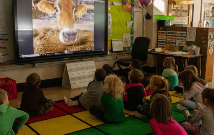 A group of children in a classroom sitting on a rug enjoying a virtual tour of a farm projected from a screen.